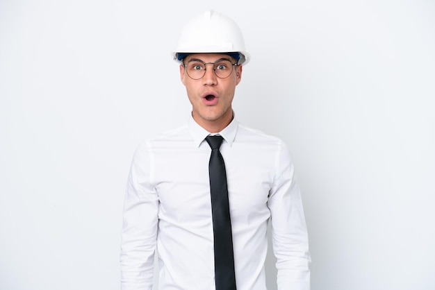 Young architect caucasian man with helmet and holding blueprints isolated on white background with surprise facial expression