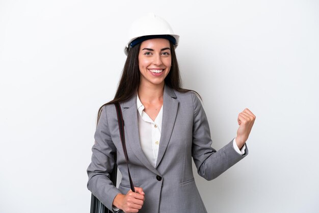 Young architect caucasian woman with helmet and holding blueprints isolated on white background pointing back