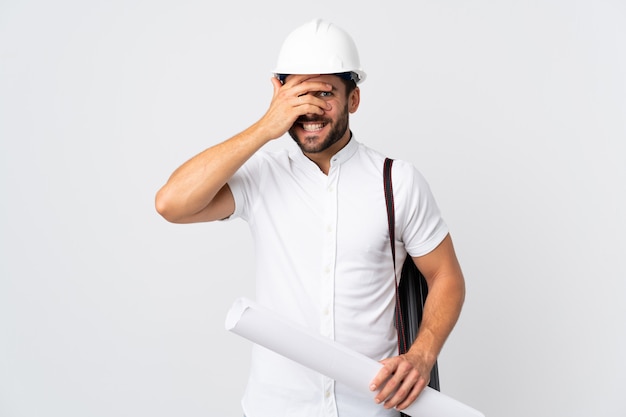 Young architect man with helmet and holding blueprints isolated on white wall covering eyes by hands and smiling