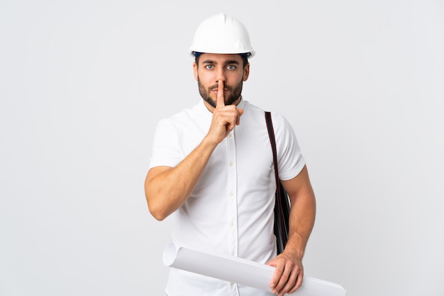 Young architect man with helmet and holding blueprints isolated on white wall showing a sign of silence gesture putting finger in mouth