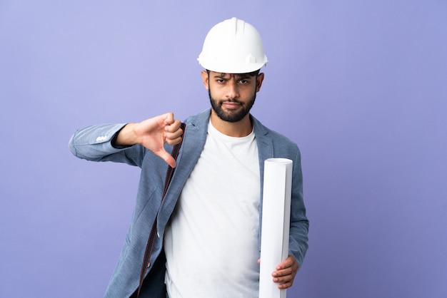 Young architect Moroccan man with helmet and holding blueprints showing thumb down with negative expression