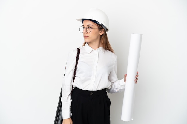 Young architect woman with helmet and holding blueprints isolated on white background looking to the side