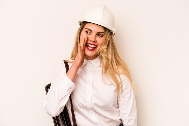 Young architect woman with helmet and holding blueprints isolated on white background shouting and holding palm near opened mouth