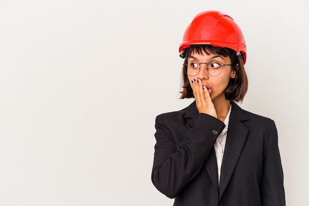 Young architect woman with red helmet isolated on white background thoughtful looking to a copy space covering mouth with hand.