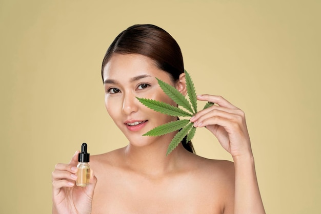 Young ardent woman with healthy skin holding green hemp leaf and cbd oil