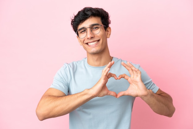 Young Argentinian man isolated on pink background With glasses making heart with hands