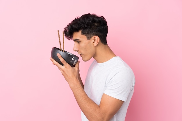 Young Argentinian man over isolated white wall holding a bowl of noodles with chopsticks and eating it