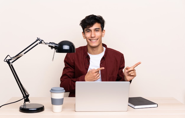 Young Argentinian man in a table with a laptop in his workplace pointing finger to the side