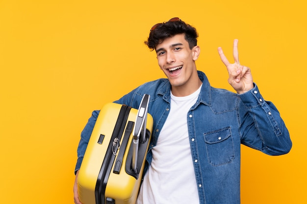 Young Argentinian man in vacation with travel suitcase and making victory gesture