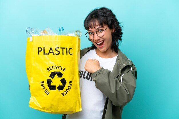 Young Argentinian woman holding a bag full of plastic bottles to recycle isolated on blue background celebrating a victory