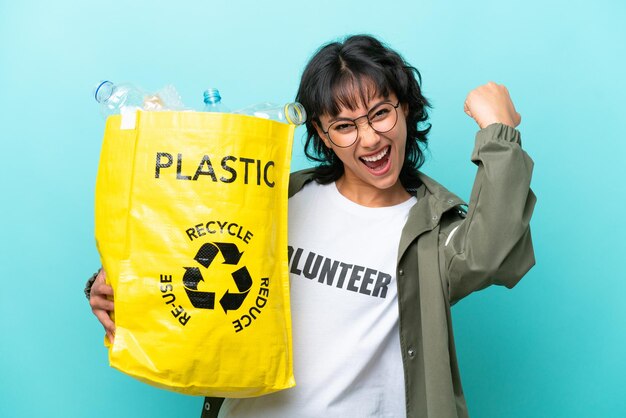 Young Argentinian woman holding a bag full of plastic bottles to recycle isolated on blue background celebrating a victory