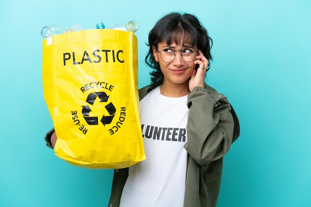 Young Argentinian woman holding a bag full of plastic bottles to recycle isolated on blue background frustrated and covering ears