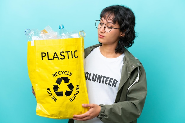 Young Argentinian woman holding a bag full of plastic bottles to recycle isolated on blue background looking to the side