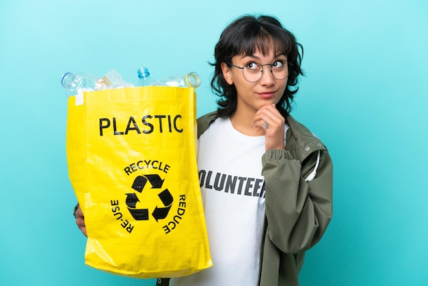 Young Argentinian woman holding a bag full of plastic bottles to recycle isolated on blue background and looking up