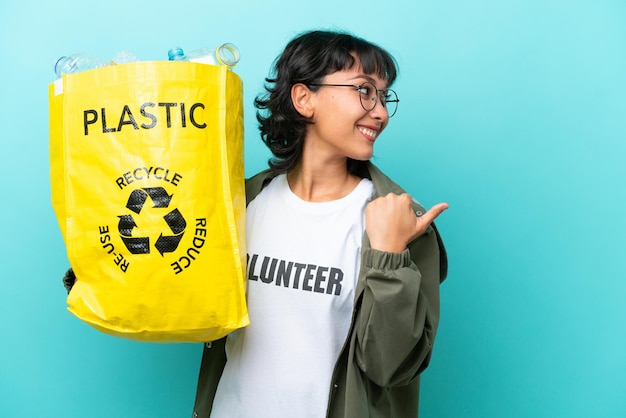 Young Argentinian woman holding a bag full of plastic bottles to recycle isolated on blue background pointing to the side to present a product