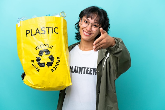 Young Argentinian woman holding a bag full of plastic bottles to recycle isolated on blue background shaking hands for closing a good deal