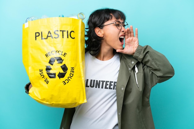 Young Argentinian woman holding a bag full of plastic bottles to recycle isolated on blue background shouting with mouth wide open to the side