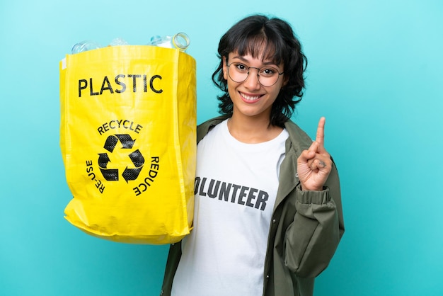 Young Argentinian woman holding a bag full of plastic bottles to recycle isolated on blue background showing and lifting a finger