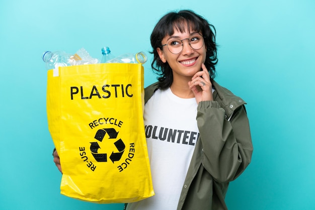 Young Argentinian woman holding a bag full of plastic bottles to recycle isolated on blue background thinking an idea while looking up