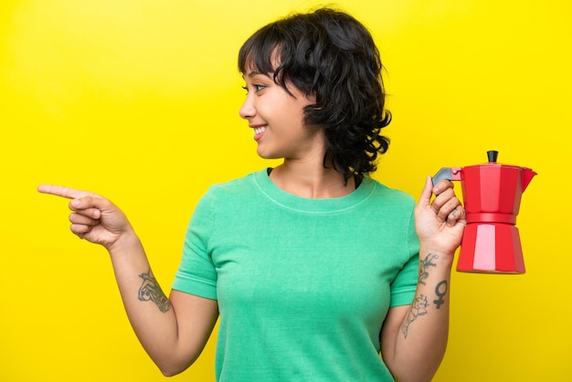 Young Argentinian woman holding coffee pot isolated on yellow background pointing to the side to present a product