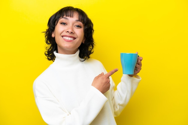 Young Argentinian woman holding cup of coffee isolated on yellow background and pointing it