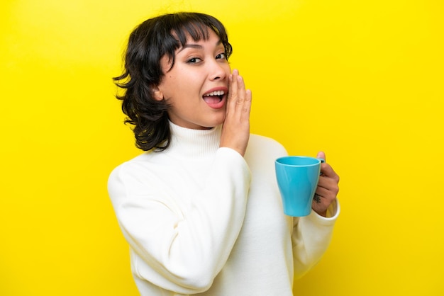 Young Argentinian woman holding cup of coffee isolated on yellow background whispering something