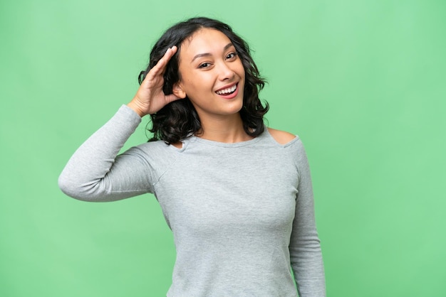 Young Argentinian woman over isolated background listening to something by putting hand on the ear