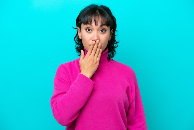 Young Argentinian woman isolated on blue background covering mouth with hand