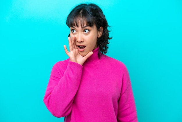 Young Argentinian woman isolated on blue background whispering something with surprise gesture while looking to the side