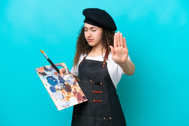 Young artist Arab woman holding a palette isolated on blue background making stop gesture and disappointed