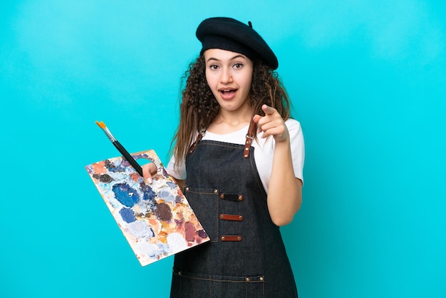 Young artist Arab woman holding a palette isolated on blue background surprised and pointing front