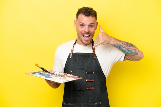 Young artist caucasian man holding a palette isolated on yellow background making phone gesture Call me back sign