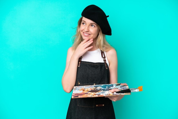 Young artist caucasian woman holding a palette isolated on blue background looking up while smiling