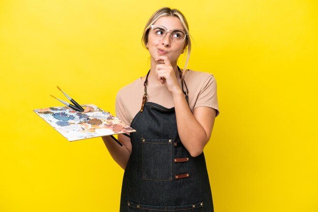 Photo young artist caucasian woman holding a palette isolated on yellow background and looking up