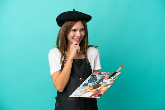 Young artist English woman holding a palette isolated on blue background looking to the side and smiling