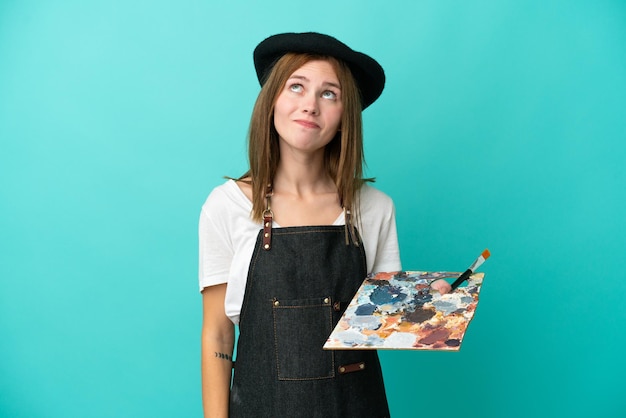 Young artist English woman holding a palette isolated on blue background and looking up