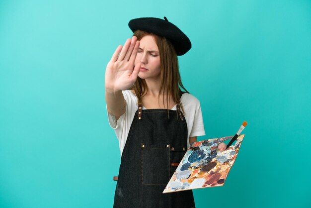 Young artist English woman holding a palette isolated on blue background making stop gesture and disappointed