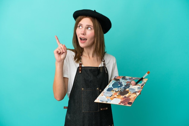 Young artist English woman holding a palette isolated on blue background thinking an idea pointing the finger up