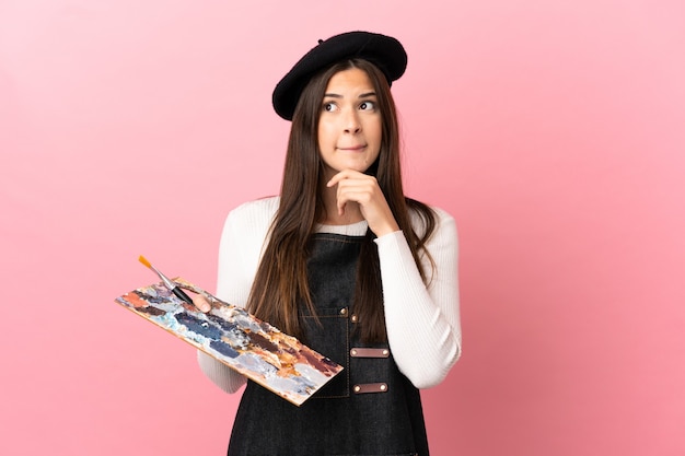 Photo young artist girl holding a palette over isolated pink background having doubts and thinking