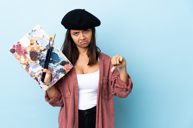 Photo young artist woman holding a palette over blue wall showing thumb down with two hands