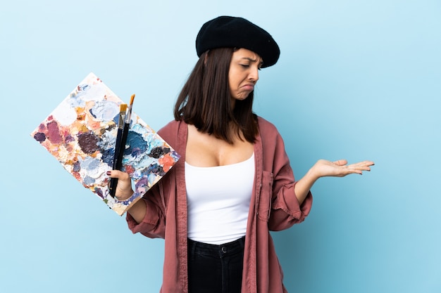 Photo young artist woman holding a palette over isolated blue holding copyspace with doubts.