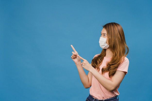 Young Asia girl wear medical face mask shows something at blank space with dressed in casual cloth and look at front isolated on blue wall