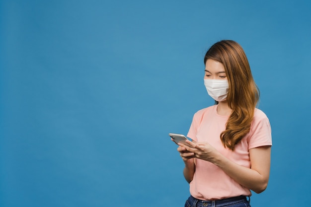 Young Asia girl wearing medical face mask using mobile phone with dressed in casual clothing isolated on blue wall