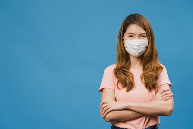 Young Asia girl wearing medical face mask with arms crossed, dressed in casual cloth and looking at front isolated on blue wall