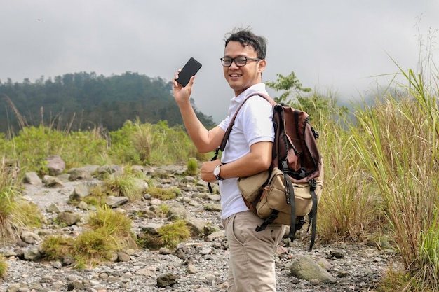 Young asian backpacker man is happy and smile when he showing smartphone when traveling in mountain