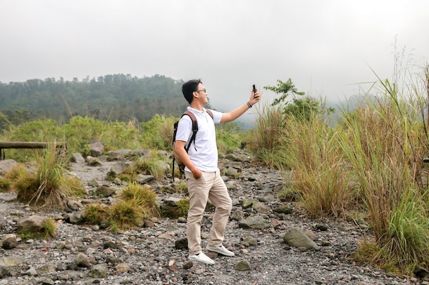 Young asian backpacker man is happy taking vlog or video with smartphone when traveling in mountain