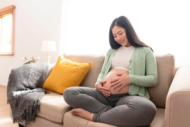 Young asian beautiful pregnant woman hands fondle on belly sittingxAon sofa at home Pregnancy maternity preparation and expectation concept