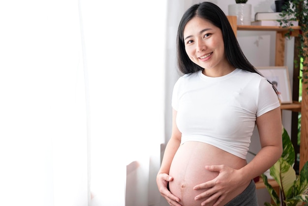 Young asian beautiful pregnant woman holds hands on belly standing near window at home Pregnancy maternity preparation and expectation concept