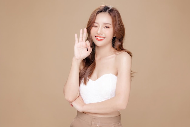 Young Asian beauty woman curly long hair with korean makeup style on face and perfect skin showing OK sign on isolated beige background Facial treatment Cosmetology plastic surgery