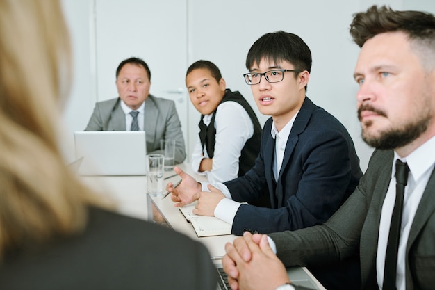 Young Asian businessman in formalwear asking business coach question while sitting between intercultural colleagues at conference